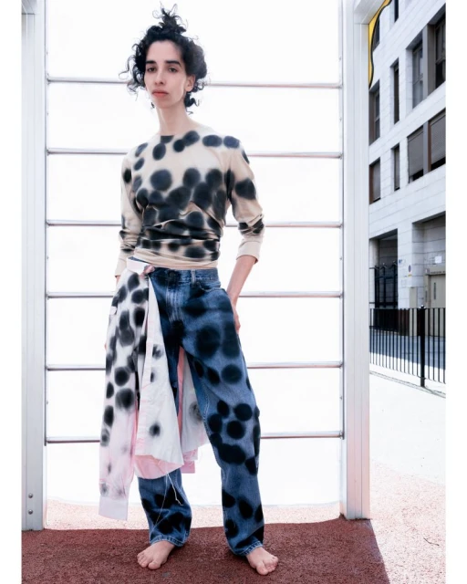 A barefoot woman stands in front of an outdoor structure wearing a polka dot shirt, dyed jeans, and a polka dot shirt tied around her waist.