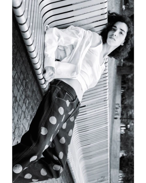 Woman lying on a modern bench wearing polka dot jeans and a white shirt in a black and white outdoor fashion shot.