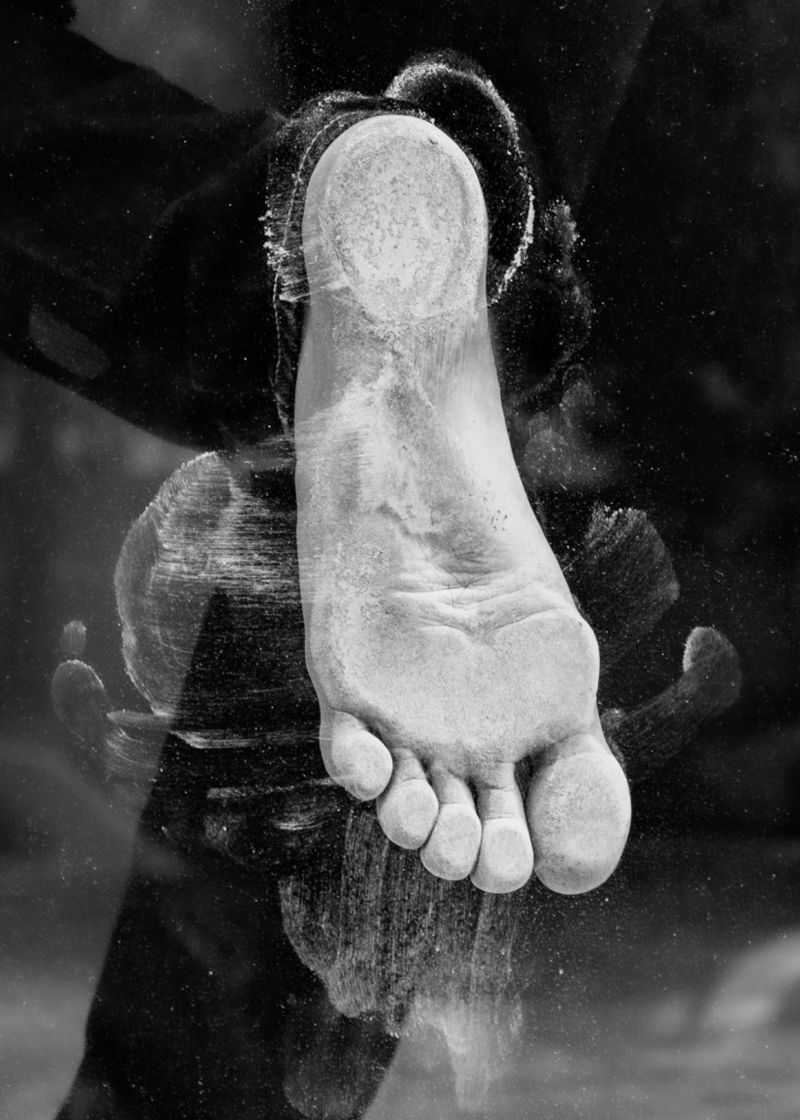 Monochrome image captures a single human foot and toes pressed against a surface, creating textured footprints, set against a dark background.