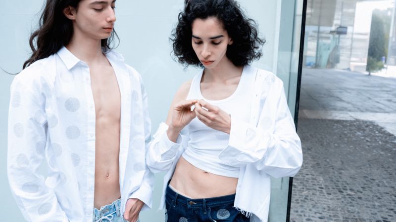 Two people pose outdoors in white button-down shirts and dark blue jeans with white polka dots standing near a glass structure.