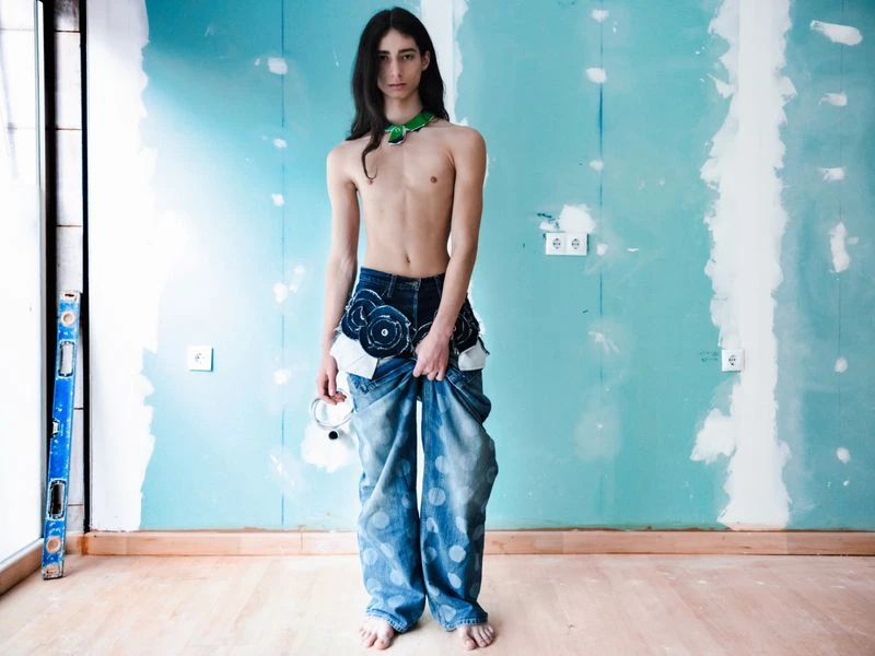 Full shot of a slender, topless adult male in baggy jeans with floral embellishments, accessorized with a green necklace in an unfinished room.