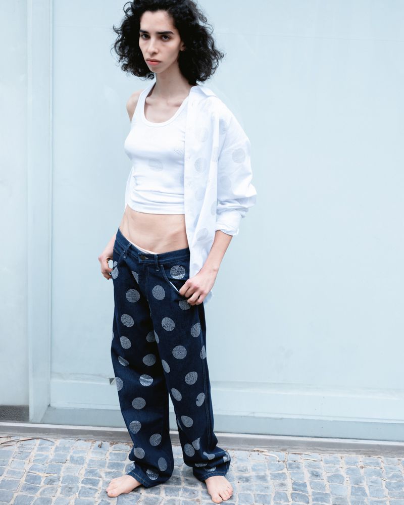 Adult female posing in a white top and polka dot jeans, standing barefoot against a light blue wall, looking at the camera.