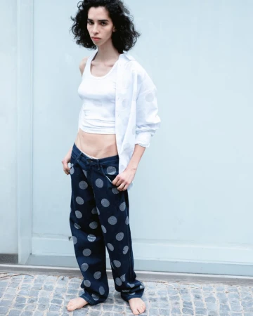 Adult female posing in a white top and polka dot jeans, standing barefoot against a light blue wall, looking at the camera.