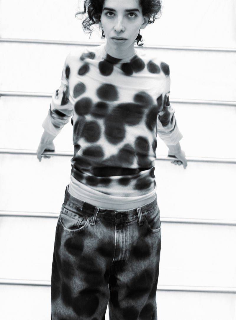 Black and white portrait of a person with curly hair wearing polka dot shirt and tie dye jeans in front of a metal staircase.