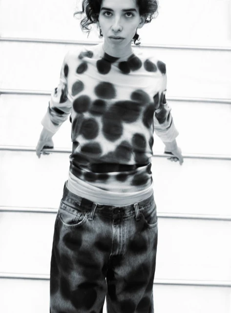 Black and white portrait of a person with curly hair wearing polka dot shirt and tie dye jeans in front of a metal staircase.