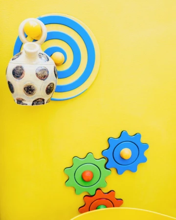 Playground equipment with colorful gears, circles, and cylinders mounted on a yellow wall with shadows, creating an engaging space.