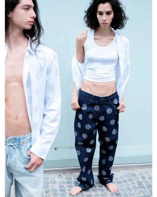 Two people pose in a studio with natural light. A man wears a white shirt and jeans; a woman wears a tank top and polka dot jeans.