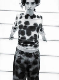 Model wearing a patterned long-sleeve top and dark denim jeans posing against horizontal rails in a studio.