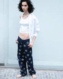 Model with dark curly hair wearing a white tank top and polka dot pants against a pale wall.