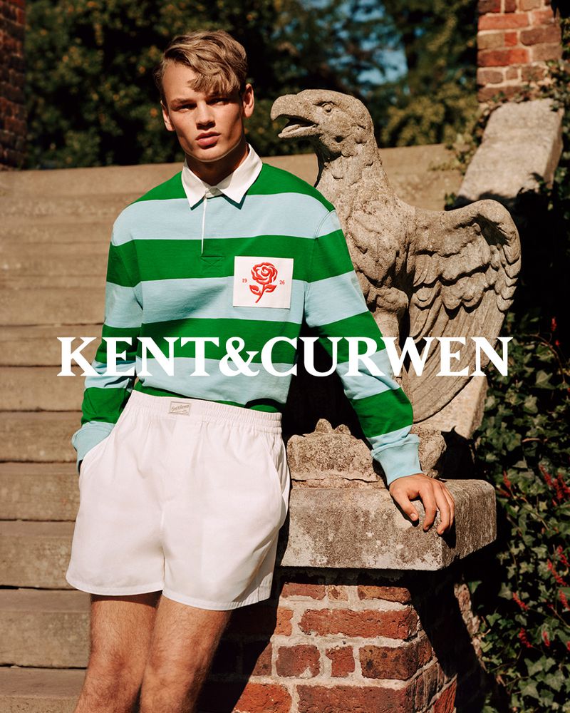 A young man in striped rugby shirt and white shorts poses next to a stone eagle sculpture on steps in front of greenery outdoors.
