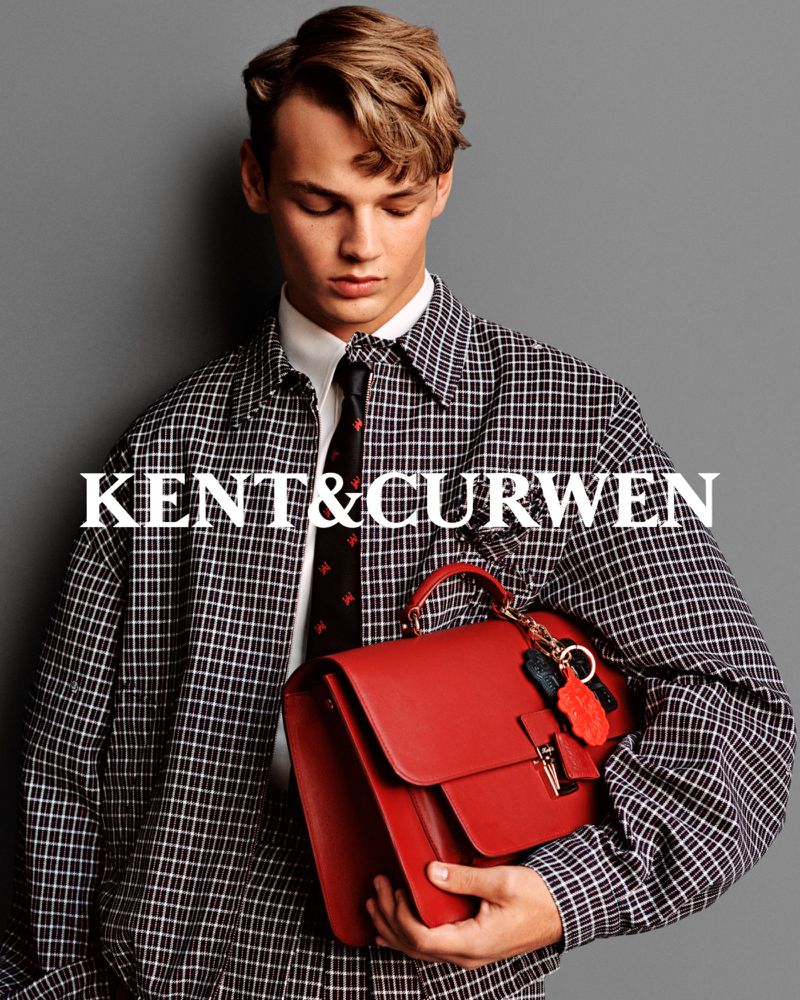 A young adult man wearing a plaid jacket and tie holds a red leather briefcase in a studio shot against a grey background.