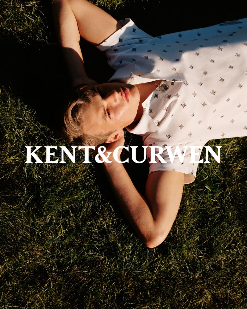 Blond male model in printed polo shirt poses lying on grass in a sunlit outdoor shot. Kent & Curwen text overlay.