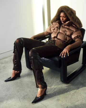 Elegant adult female seated in chair wearing brown puffer coat with fur hood, matching pants and heels against a light-grey indoor backdrop.