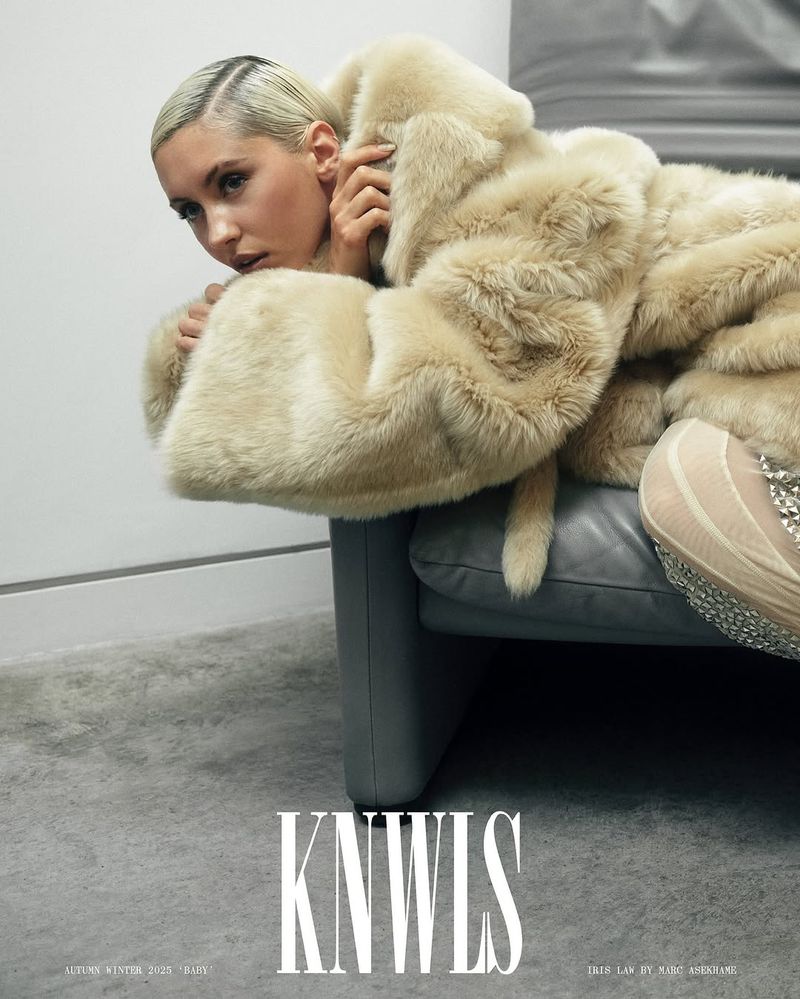 Blonde woman in a fur coat poses on a gray couch in a studio setting, fashion photography, neutral tones, soft lighting, looking at the camera.