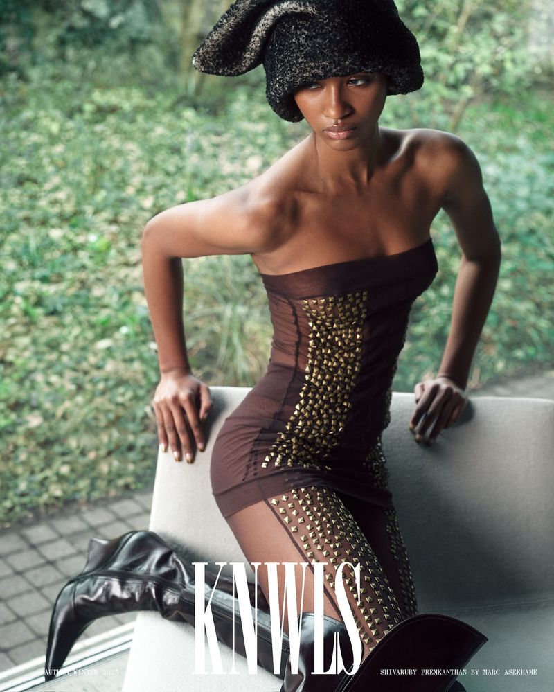 An adult female fashion model wearing a strapless studded dress, knee-high boots, and a hat poses near a window with a blurred green background.