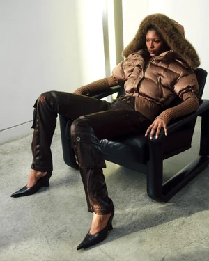 Model seated in a black armchair wearing a brown puffer jacket and metallic trousers against a neutral wall