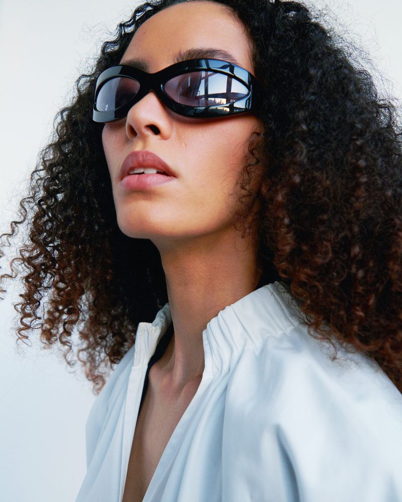 Woman with curly hair wears sunglasses and a light blue blouse in a studio portrait, against a white backdrop.