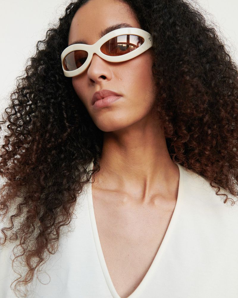 Woman wearing cream-colored v-neck blouse and sunglasses with dark curly hair in a close-up shot against a white backdrop.