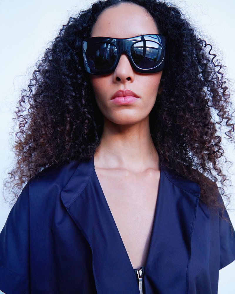Close-up of an adult female with curly hair wearing oversized sunglasses and a navy-blue jacket against a white backdrop.