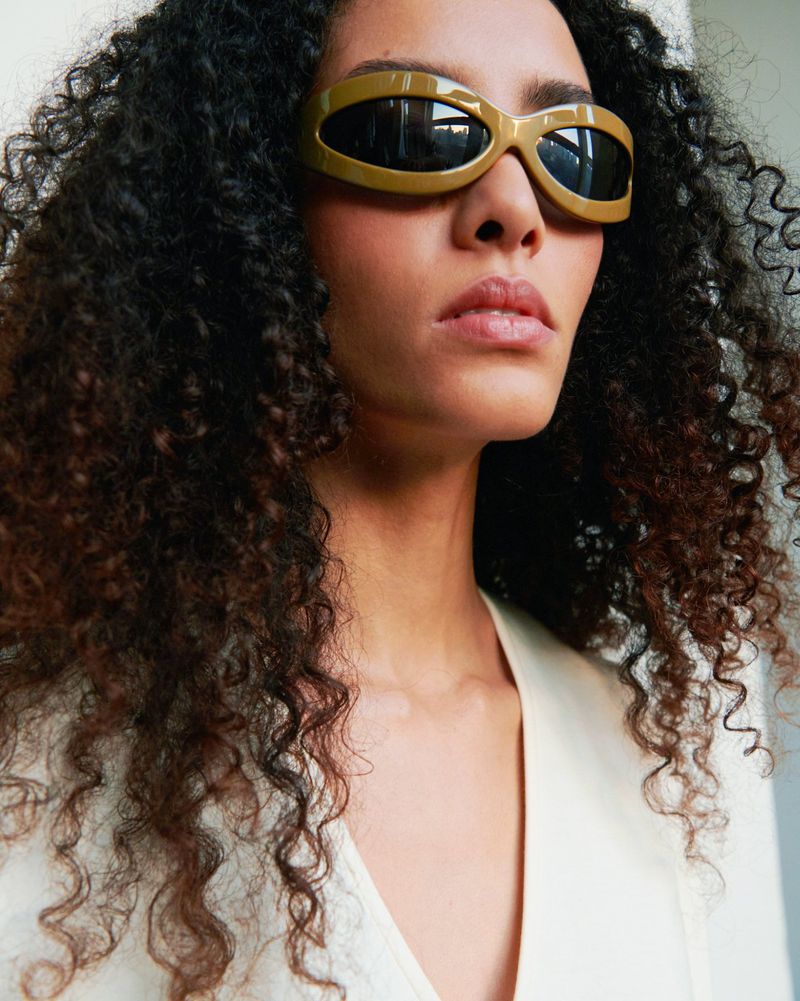 Portrait of a woman with curly hair wearing light-brown sunglasses and a light-colored shirt against a neutral background.