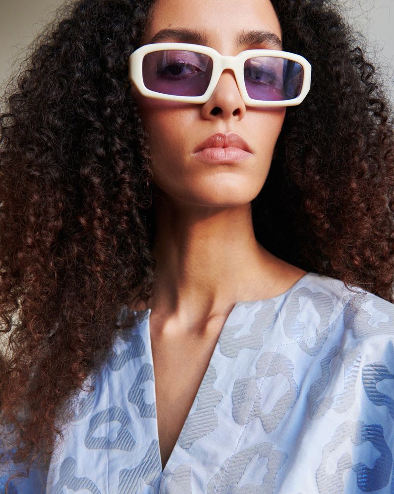 Headshot of a woman with curly hair wearing stylish sunglasses and a patterned top, posing in a studio with soft lighting.
