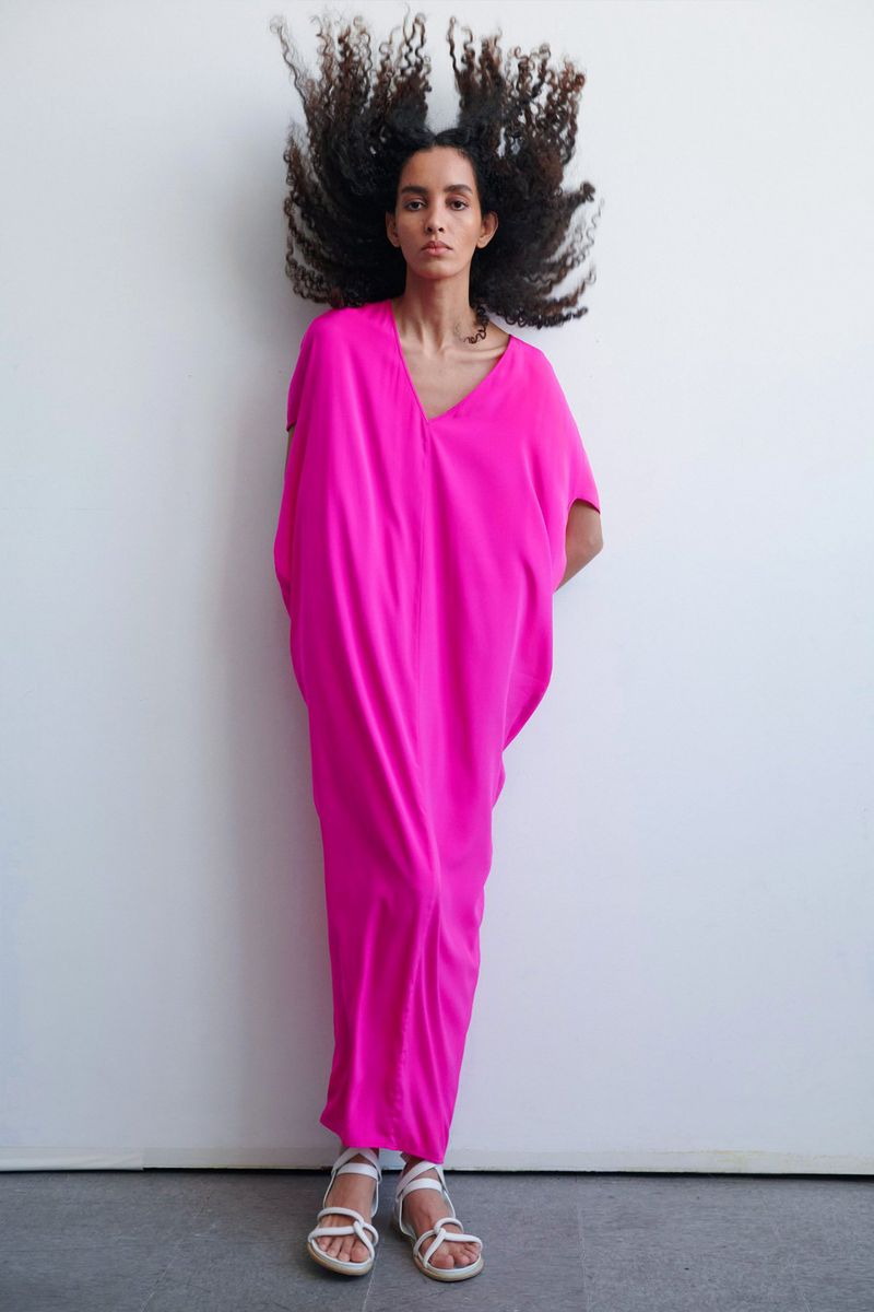 Fashionable woman in a hot pink dress stands against a white wall, her afro blowing upward, and wearing strappy white sandals.