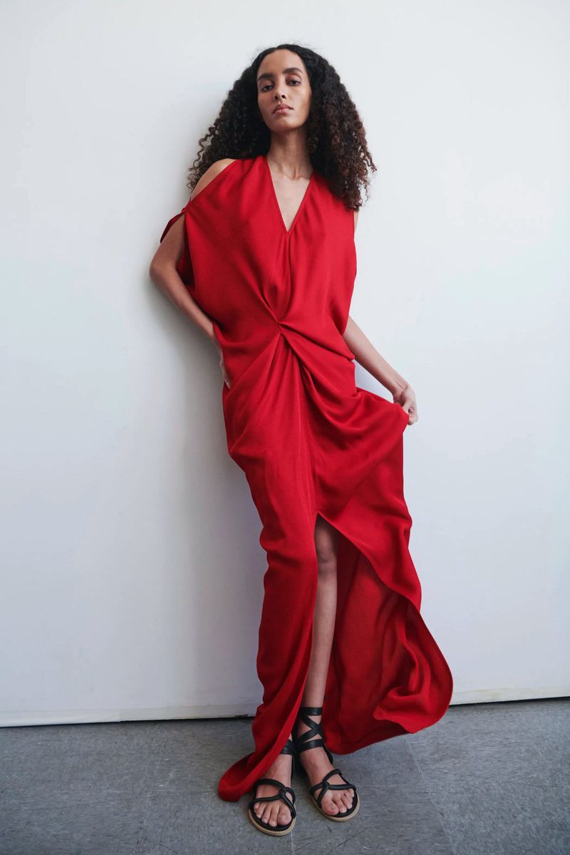 A woman poses in a studio wearing a red dress with black strappy sandals, leaning against a white wall with a neutral facial expression.