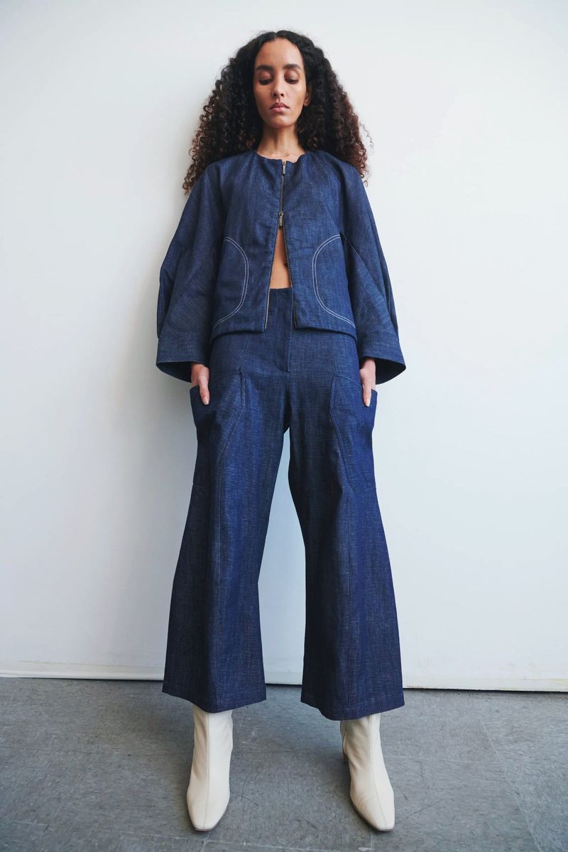 Model with long dark curly hair wearing a blue denim jacket and matching trousers with light boots against a white wall