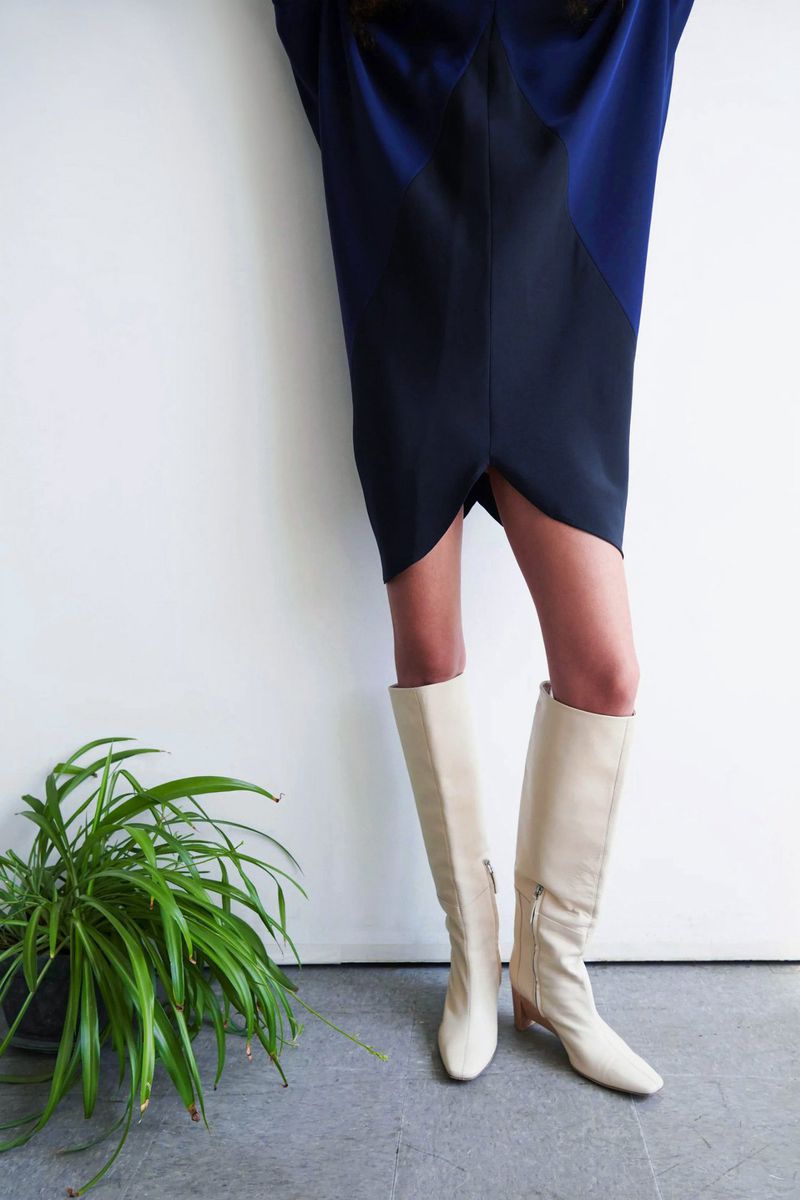 Fashionable knee-high cream-colored leather boots paired with a stylish navy blue dress, accompanied by a vibrant green houseplant.