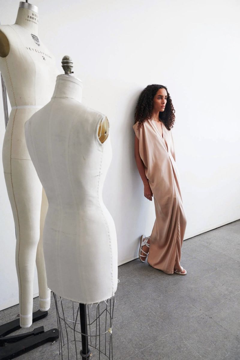 A woman in a dress poses next to two dress forms against a white wall, with light and airy studio lighting, in a vertical portrait.