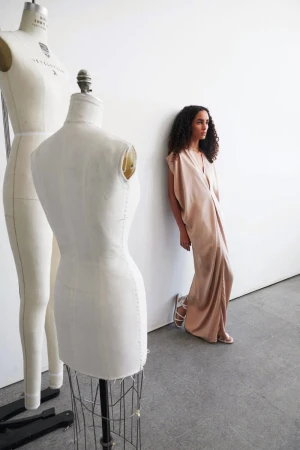 Model standing next to dressmaker's dummies wearing a long nude dress against a plain white wall
