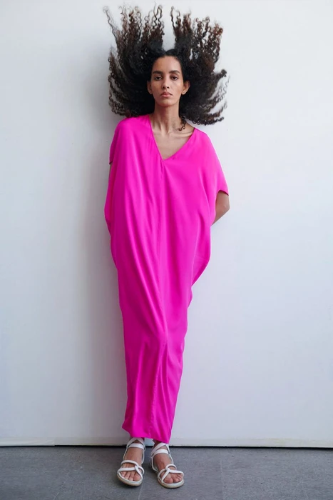 Model in vibrant pink dress with voluminous dark curly hair standing against white wall