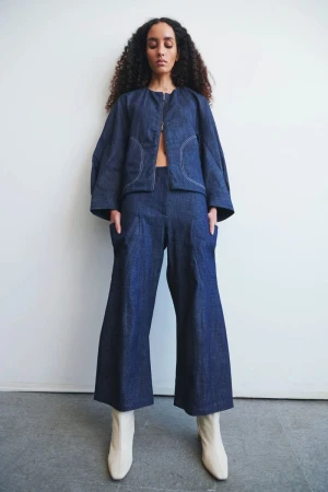 Model with long dark curly hair wearing a blue denim jacket and matching trousers with light boots against a white wall