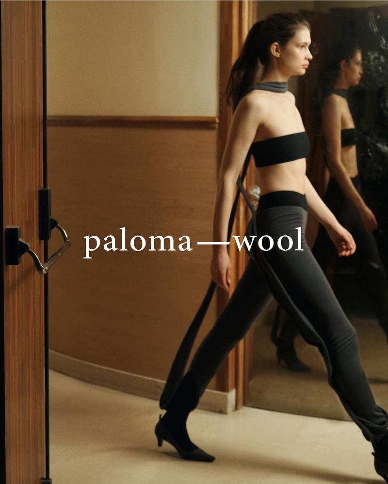 A woman in a dark bra top and leggings walks past a mirror in an indoor hallway, creating a double reflection and emphasizing her style.