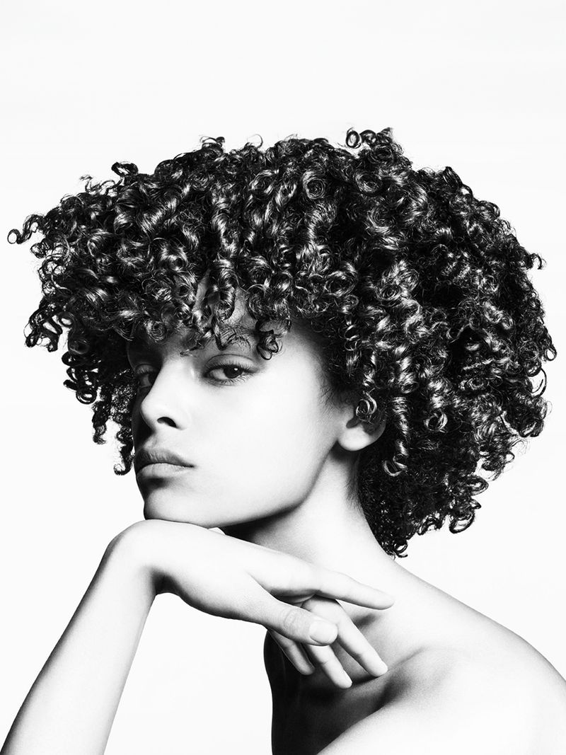 Monochromatic portrait of a curly-haired young woman with hand to chin, bare shoulders, and neutral facial expression against a white background.