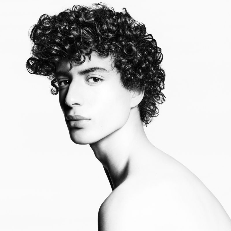 Striking black and white portrait of a young adult male with curly dark hair, gazing intently at the camera against a clean white backdrop.
