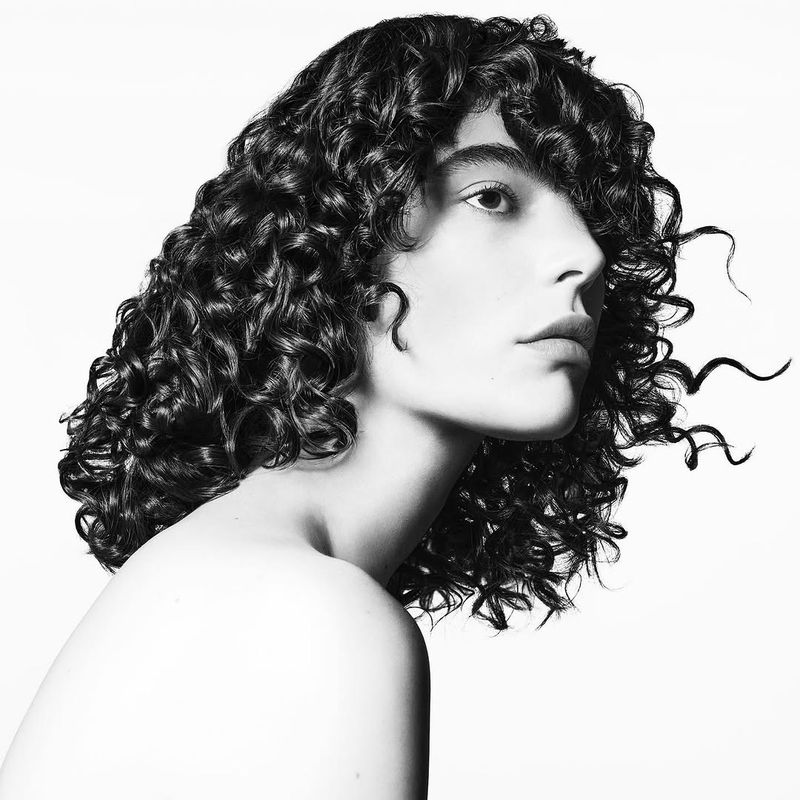 Monochrome portrait of a young person with curly hair, captured in a studio setting with a clean, white backdrop and soft lighting.