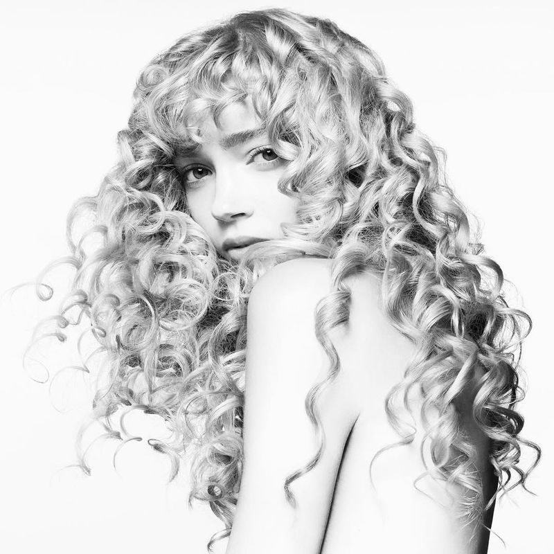 Monochrome portrait of a young woman with blonde, curly hair looking over her shoulder against a bright white studio backdrop.