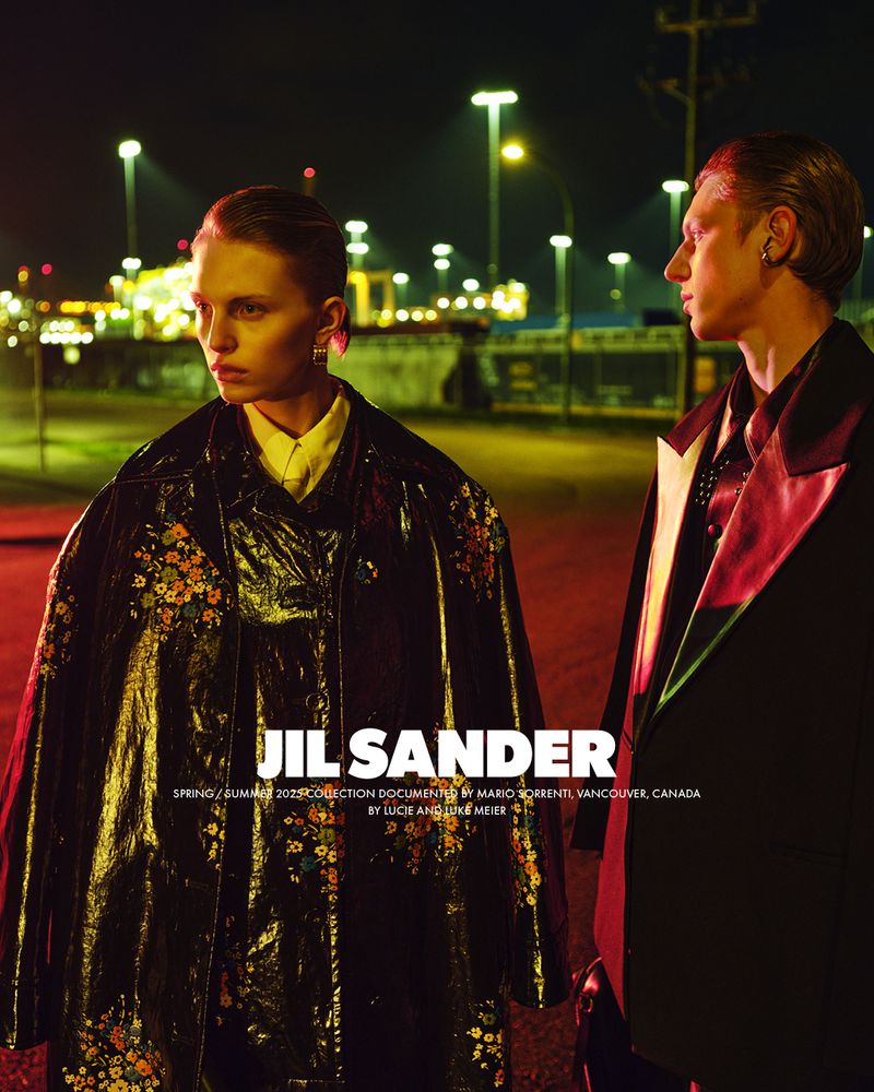 Fashion shot of two models wearing dark coats with floral pattern in urban setting at night with street lights and architectural elements.