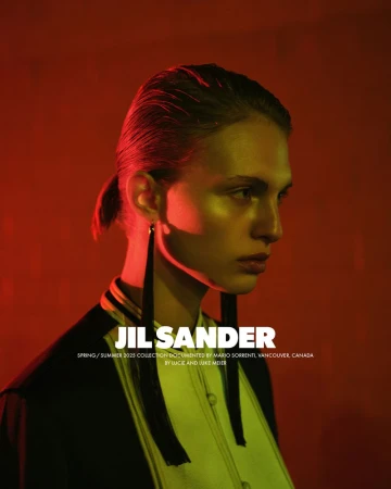 A model showcases Jil Sander's spring/summer collection in a studio setting, illuminated by striking red and yellow lighting, wearing long black earrings.