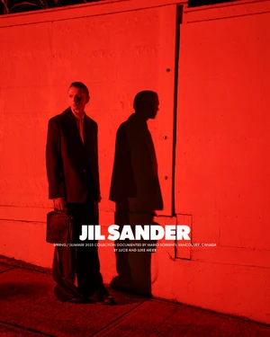 Model wearing dark blazer standing against a red-lit wall casting a prominent shadow for Jil Sander