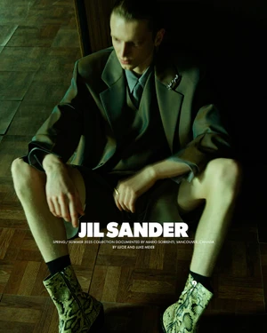 Model seated on a wooden floor wearing an olive suit jacket and patterned boots for the Jil Sander SS25 campaign.