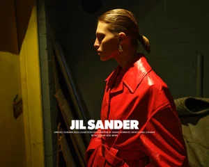 Model with tied-back hair and statement earring wearing a glossy red belted coat in a dim interior setting for Jil Sander S/S 2025 campaign.