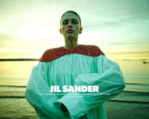Model wearing a light blue dress with red embellished shoulders against a coastal sunset background for Jil Sander S/S 2025 campaign.