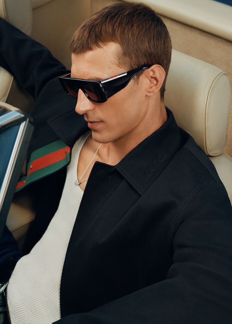 A fashionable adult male model wears sunglasses and a black jacket in a close-up, angled shot, showcasing fashion and style.