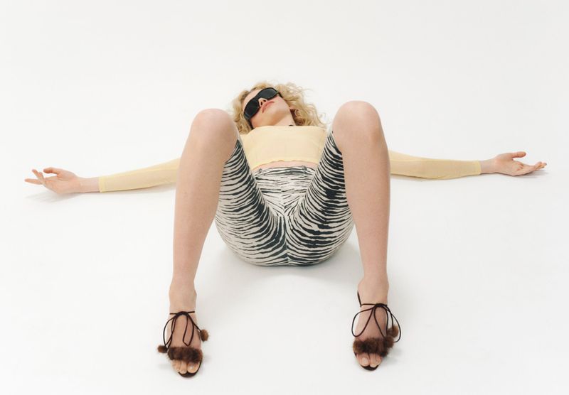 A woman lies on a white background in a yellow top, zebra-print leggings, and brown heeled sandals, with her arms outstretched and wearing sunglasses.