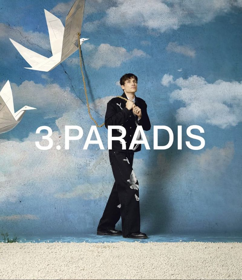 Man in black jacket and pants stands against blue sky background with paper birds. Fashion editorial shot. 3.PARADIS logo.
