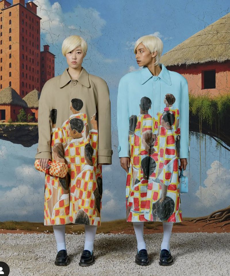 Two fashion models in patterned trench coats stand against a painted backdrop of a building and hut on floating ground.