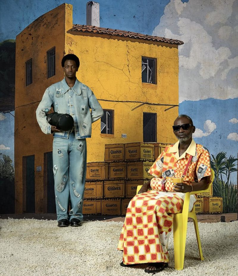Two men pose in front of a painted backdrop featuring a yellow building, blue sky, and Guiness crates, captured with a full shot.