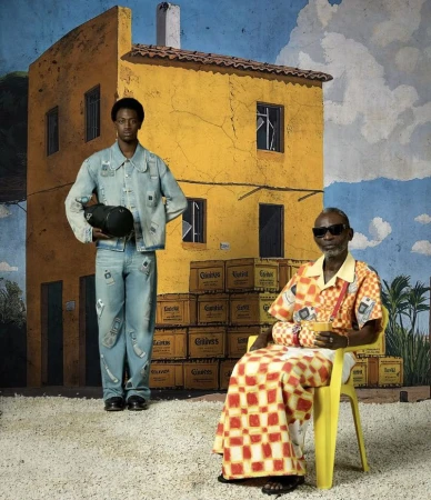 Two men pose in front of a painted backdrop featuring a yellow building, blue sky, and Guiness crates, captured with a full shot.